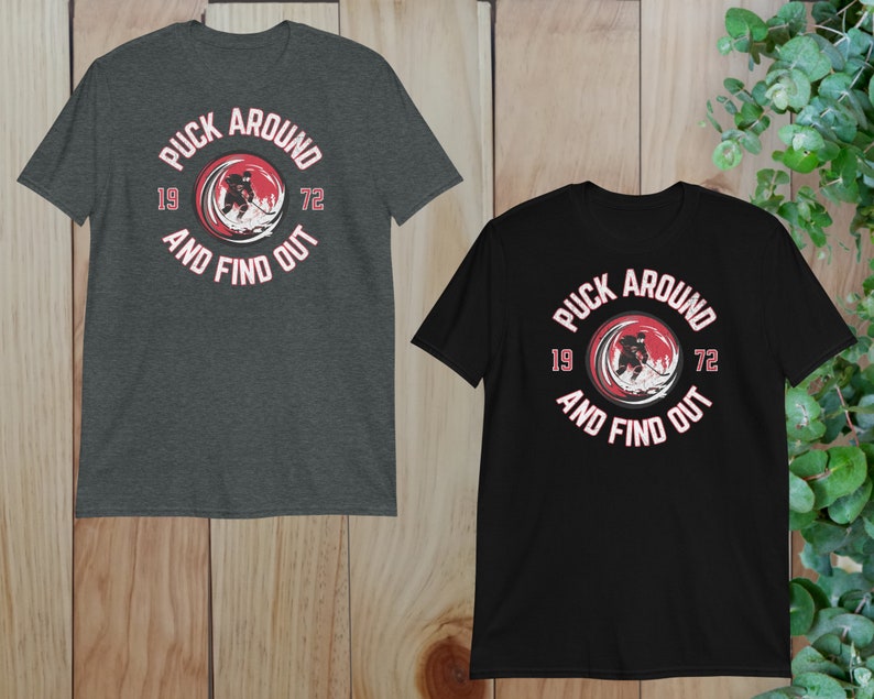 May include: Two t-shirts with a hockey player graphic and the text "Puck Around And Find Out" in a red circle with white text. The numbers 19 and 72 are on either side of the circle. One shirt is dark heather grey and the other is black.