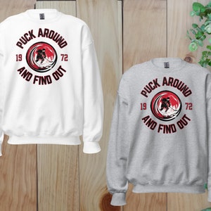 May include: Two sweatshirts with a hockey player graphic and the text "Puck Around And Find Out" in a red circle with black text. The sweatshirt on the left is white and the sweatshirt on the right is gray.