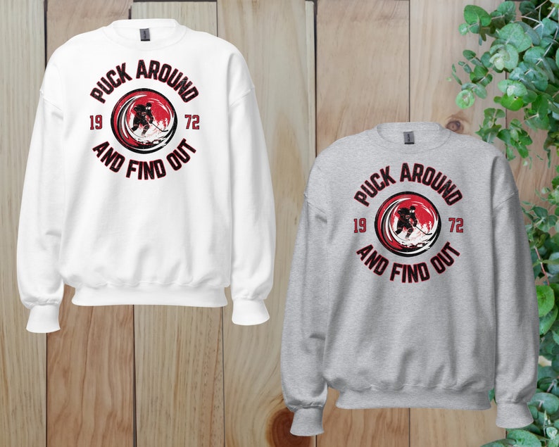 May include: Two sweatshirts with a hockey player graphic and the text "Puck Around And Find Out" in a red circle with black text. The sweatshirt on the left is white and the sweatshirt on the right is gray.