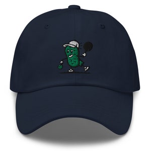 May include: A navy blue baseball cap with an embroidered green pickle character holding a tennis racket. The pickle is wearing a white hat and has a smiling face.