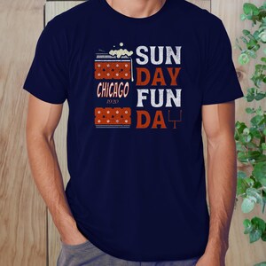 May include: Navy blue t-shirt with the words "SUNDAY FUN DAY" in white and orange block letters. The shirt also features the text "CHICAGO 1920" and a football goal post graphic. The design includes a beer mug graphic.