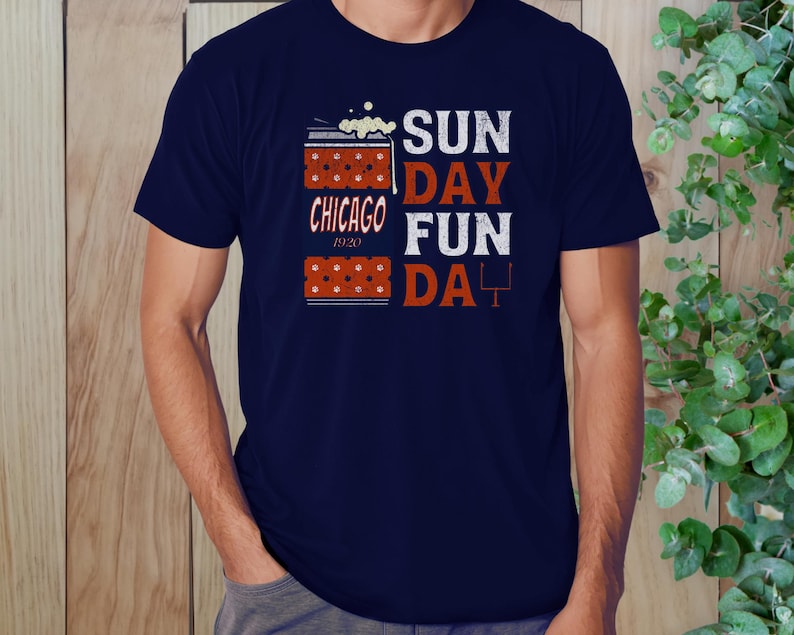 May include: Navy blue t-shirt with the words "SUNDAY FUN DAY" in white and orange block letters. The shirt also features the text "CHICAGO 1920" and a football goal post graphic. The design includes a beer mug graphic.