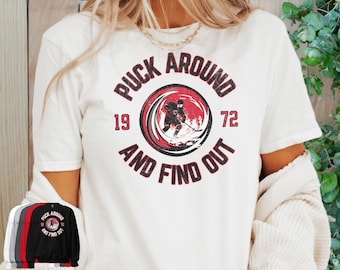 Puede incluir: Camiseta blanca con un gráfico rojo y negro que dice "Puck Around and Find Out" con un jugador de hockey en el centro. Los números 19 y 72 están a cada lado del gráfico.