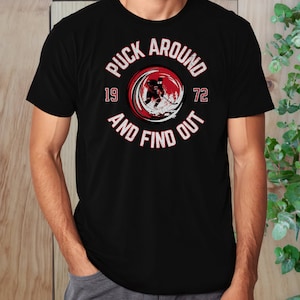 May include: Black t-shirt with a red circle graphic featuring a hockey player and the text "Puck Around 19 72 And Find Out".
