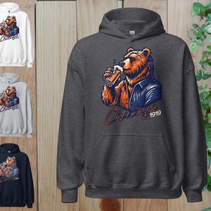 May include: A dark grey hooded sweatshirt featuring a cartoon illustration of a bear wearing a blue jacket and holding a beer. The bear is looking to the right and the text "Chicago 1919" is printed below the bear.
