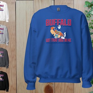 May include: A blue sweatshirt with the word "BUFFALO" in red block letters above a cartoon hot dog character. The text "GOT THAT DOG IN ME" is below the hot dog. Additional sweatshirts in white, gray, dark gray, and black are shown.