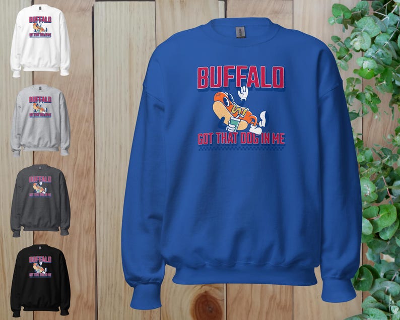 May include: A blue sweatshirt with the word "BUFFALO" in red block letters above a cartoon hot dog character. The text "GOT THAT DOG IN ME" is below the hot dog. Additional sweatshirts in white, gray, dark gray, and black are shown.