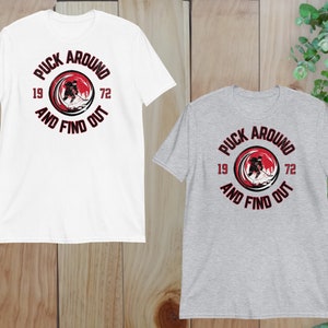 May include: Two t-shirts with a hockey-themed graphic. The graphic features a hockey player skating with the text "Puck Around and Find Out" in a circle around the player. The text "19" and "72" are on either side of the circle. The shirt on the left is white and the shirt on the right is gray.