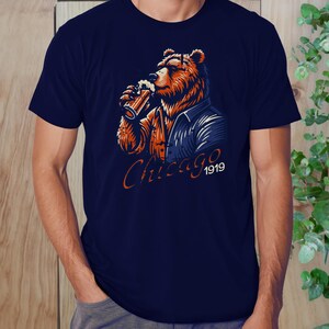 May include: A navy blue t-shirt featuring a cartoon illustration of a bear wearing a blue jacket and holding a glass of beer. The text "Chicago 1919" is printed below the bear.