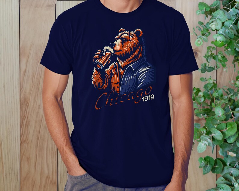 May include: A navy blue t-shirt featuring a cartoon illustration of a bear wearing a blue jacket and holding a glass of beer. The text "Chicago 1919" is printed below the bear.