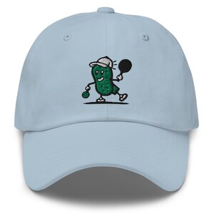 May include: A light blue baseball cap with an embroidered green pickle character wearing a white hat and holding a tennis racket. The pickle character is smiling and has a green ball in its hand.