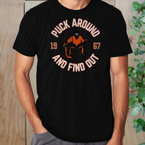May include: Black t-shirt with a white graphic of a hockey goalie in an orange uniform with the text "Puck Around 19 67 And Find Out".