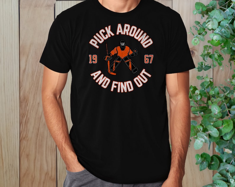 May include: Black t-shirt with a white graphic of a hockey goalie in an orange uniform with the text "Puck Around 19 67 And Find Out".