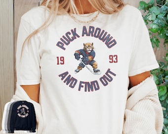 May include: White t-shirt with a vintage-style graphic of a cartoon hockey player with the text "Puck Around and Find Out" and the numbers "19" and "93".