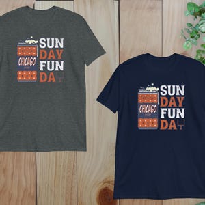 May include: Two short-sleeved t-shirts, one charcoal gray and one navy blue, are displayed. Each shirt features the text "SUNDAY FUN DAY" in white and orange, with "CHICAGO 2020" and a football graphic. The shirts are on a wooden surface.