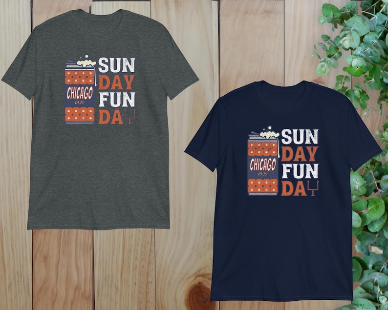 May include: Two short-sleeved t-shirts, one charcoal gray and one navy blue, are displayed. Each shirt features the text "SUNDAY FUN DAY" in white and orange, with "CHICAGO 2020" and a football graphic. The shirts are on a wooden surface.