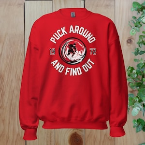 May include: Red sweatshirt with a white graphic of a hockey player in a circle with the text "Puck Around 19 72 And Find Out".