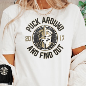 Op de afbeelding: Wit T-shirt met een zwarte en gouden afbeelding met de tekst "Puck Around and Find Out" met een ridderhelm in het midden. De nummers 20 en 17 staan ​​aan weerszijden van de helm.