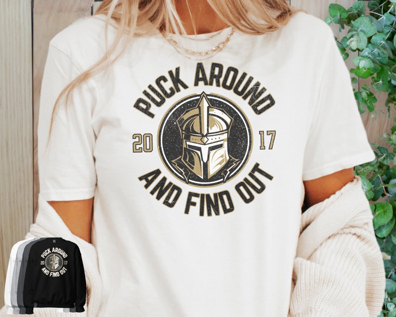 Op de afbeelding: Wit T-shirt met een zwarte en gouden afbeelding met de tekst "Puck Around and Find Out" met een ridderhelm in het midden. De nummers 20 en 17 staan ​​aan weerszijden van de helm.