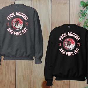 May include: Two sweatshirts with a hockey player graphic and the text "Puck Around 19 72 And Find Out". One sweatshirt is black and the other is dark gray.
