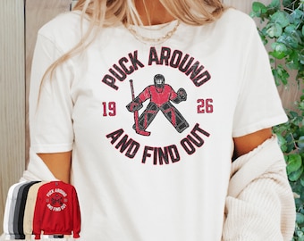 Może przedstawiać: Biała koszulka z czerwonym kołem graficznym przedstawiającym hokeistę bramkarza z tekstem "Puck Around and Find Out" i numerami "19" i "26".