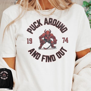 May include: White t-shirt with a red and black graphic of a hockey player with the text "Puck Around and Find Out" and the numbers "19" and "74".