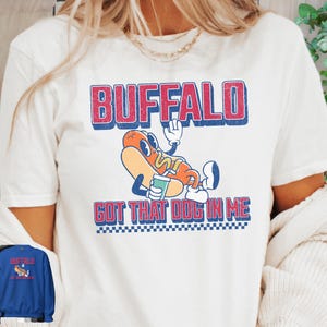 May include: White t-shirt featuring a cartoon hot dog character with the text "BUFFALO GOT THAT DOG IN ME" in red and blue. The hot dog is wearing a hat and holding a drink. A blue sweatshirt with the same design is also visible.