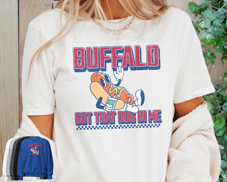 May include: White t-shirt featuring a cartoon hot dog character with the text "BUFFALO GOT THAT DOG IN ME" in red and blue. The hot dog is wearing a hat and holding a drink. A blue sweatshirt with the same design is also visible.
