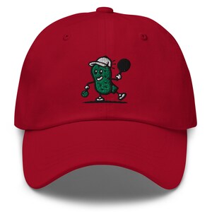 May include: Red baseball cap with an embroidered green pickle character holding a tennis racket. The pickle is wearing a white hat and has a smiling face.