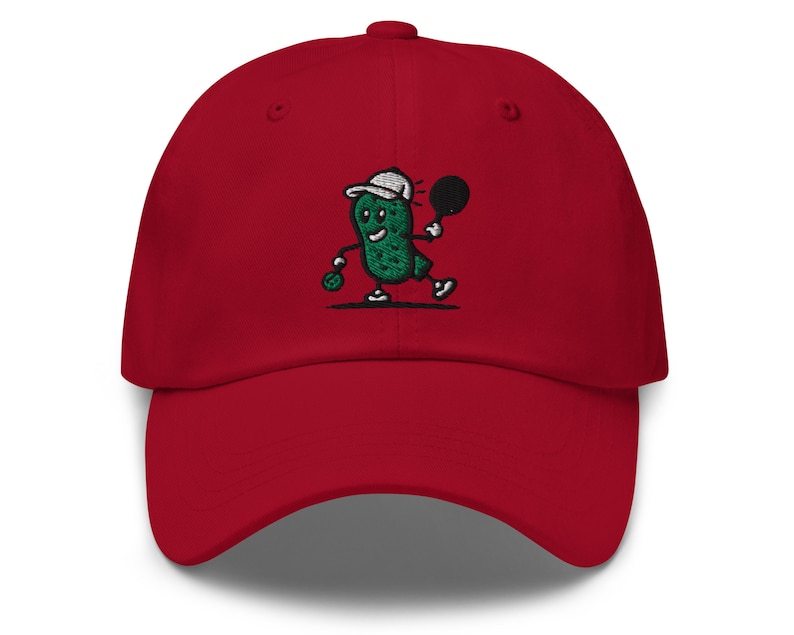 May include: Red baseball cap with an embroidered green pickle character holding a tennis racket. The pickle is wearing a white hat and has a smiling face.