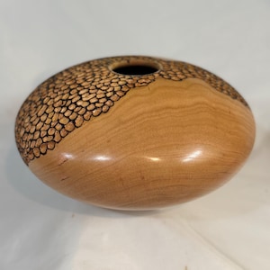 May include: A rounded wooden vessel with a smooth, light brown natural wood grain finish. The upper portion features a dark, textured, burnt-in pattern of small, irregular, pebble-like shapes that contrast with the plain, polished wood surface.