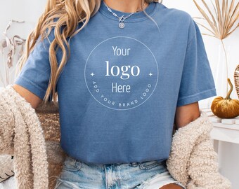Op de afbeelding: Een blauw jeanskleurig t-shirt met korte mouwen en een witte cirkelvormige afbeelding op de borst. De afbeelding bevat in het midden de tekst "Your logo Here", omringd door de woorden "ADD YOUR BRAND LOGO" en twee kleine vierpuntige sterren.
