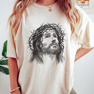 May include: Ivory-colored t-shirt featuring a detailed black and white illustration of a man wearing a crown of thorns. The graphic is centered on the shirt. The text "Ivory Color" is at the bottom right.
