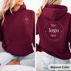 May include: Maroon-colored hoodie with a front pocket and hood. The front and back of the hoodie feature a circular design with the text "Your logo Here". The bottom right corner of the image has the text "Maroon Color".