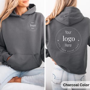 May include: Charcoal-colored hoodie with a front pocket and a hood. The front and back of the hoodie feature a circular design with the text "Your logo Here" and "Add your brand logo". The hoodie is a solid color.