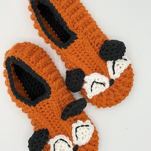 May include: A pair of orange crocheted slippers with black and white fox faces. The slippers have black trim around the opening.