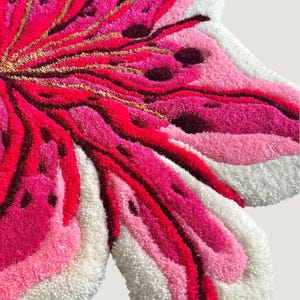 May include: A tufted rug shaped like a flower with layered petals in shades of hot pink, light pink, and white. The design features dark maroon accents and thin gold-colored lines radiating from the center, creating a textured, plush surface.