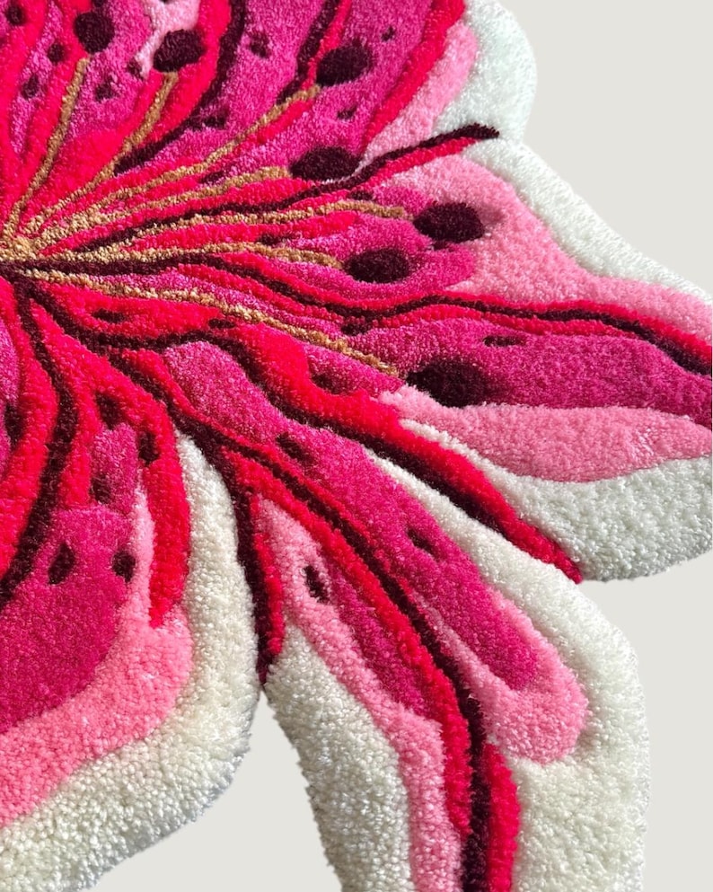 May include: A tufted rug shaped like a flower with layered petals in shades of hot pink, light pink, and white. The design features dark maroon accents and thin gold-colored lines radiating from the center, creating a textured, plush surface.