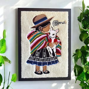 May include: Framed textile art showing a person in traditional Peruvian dress holding a white llama. The artwork features a brown hat, a colourful striped shawl, and a black skirt with blue trim. The word "perú" is written in cursive.