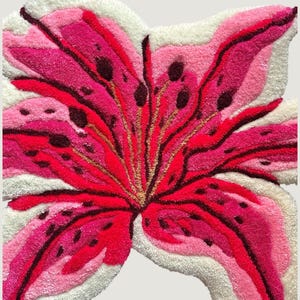May include: A tufted rug shaped like a large lily flower with vibrant pink, magenta, and deep red petals featuring dark brown spots. The flower design is outlined and accented with white tufted fibers, creating a textured, floral-shaped floor piece.
