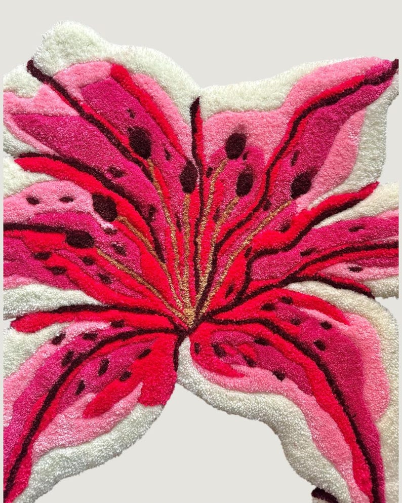 May include: A tufted rug shaped like a large lily flower with vibrant pink, magenta, and deep red petals featuring dark brown spots. The flower design is outlined and accented with white tufted fibers, creating a textured, floral-shaped floor piece.