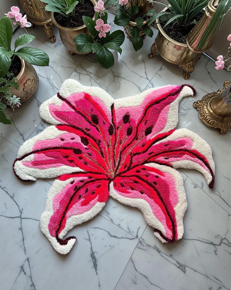 May include: A tufted rug shaped like a large lily flower with six petals. The design features vibrant shades of hot pink, magenta, and deep red, with dark brown spots and white edges. The texture is thick and plush, mimicking the organic form of a lily.
