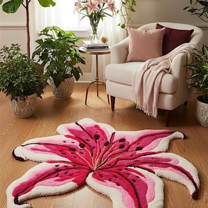 May include: A tufted area rug shaped like a large lily flower, featuring vibrant shades of deep magenta, light pink, and white. The rug has a textured, plush surface with detailed petal contours and dark spots, mimicking the appearance of a blooming lily.