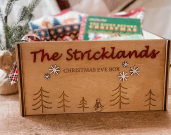 Op de afbeelding: Houten kerstavonddoos met de naam "The Stricklands" in rode letters. De doos is versierd met sneeuwvlokken, kerstbomen en een sneeuwpop. De doos is gevuld met cadeaus, waaronder een boek en cadeaupapier met patronen.