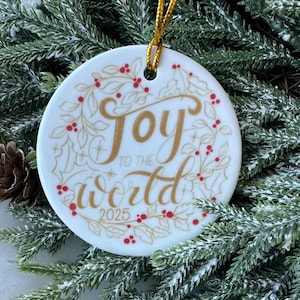 Op de afbeelding: Een witte keramische kerstbal met de gouden tekst "Joy to the World", omringd door een krans van gouden bladeren en rode bessen. Het jaar "2025" staat onderaan gedrukt. De bal hangt aan een gouden koord en ligt op dennentakken.