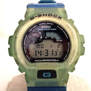 Puede incluir: Un reloj digital G-Shock con una caja verde translúcida y detalles azules. La esfera del reloj muestra la hora, la fecha y otras funciones. El reloj tiene el texto "G-SHOCK" y "CASIO" en la esfera.