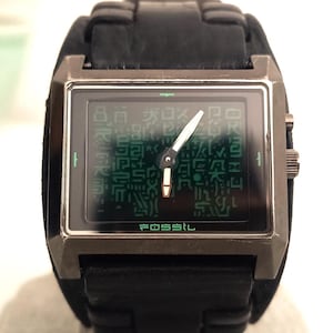 May include: A black Fossil watch with a rectangular face and a black leather band. The watch face displays a green digital display with a unique pattern. The word "FOSSIL" is printed at the bottom of the watch face.