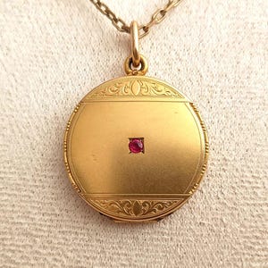 May include: A round gold locket pendant suspended from a matching gold chain. The locket features a smooth, matte gold finish with a small, square-cut red gemstone set in the centre. The top and bottom edges are decorated with intricate engraved scrollwork.