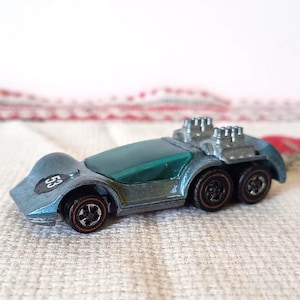 Op de afbeelding: Een vintage speelgoedauto van gegoten metaal in blauw en zilver, met zes wielen en een groene voorruit. De auto heeft het nummer "53" op de zijkant en banden met rode randen.