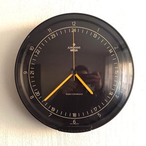 Puede incluir: Un reloj de pared negro Junghans Mega radiocontrolado. La esfera tiene números y marcadores blancos, con manecillas amarillas. El reloj está montado en una pared blanca. El texto "JUNGHANS MEGA" y "RADIO CONTROLLED" son visibles.